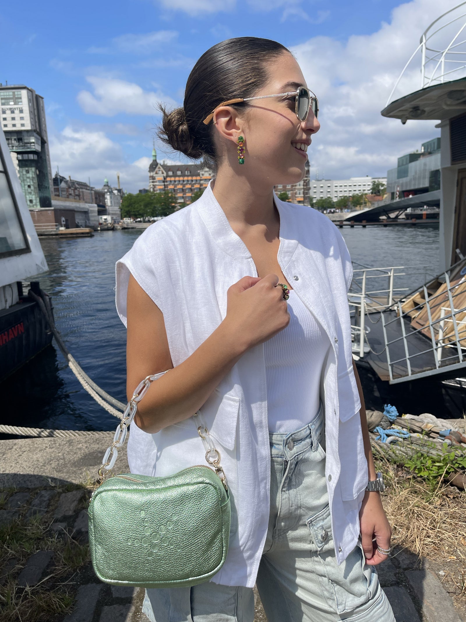 Close up Person standing by a waterfront with a Green Metallic Grabriela handbag on hand, boats, and buildings in the background