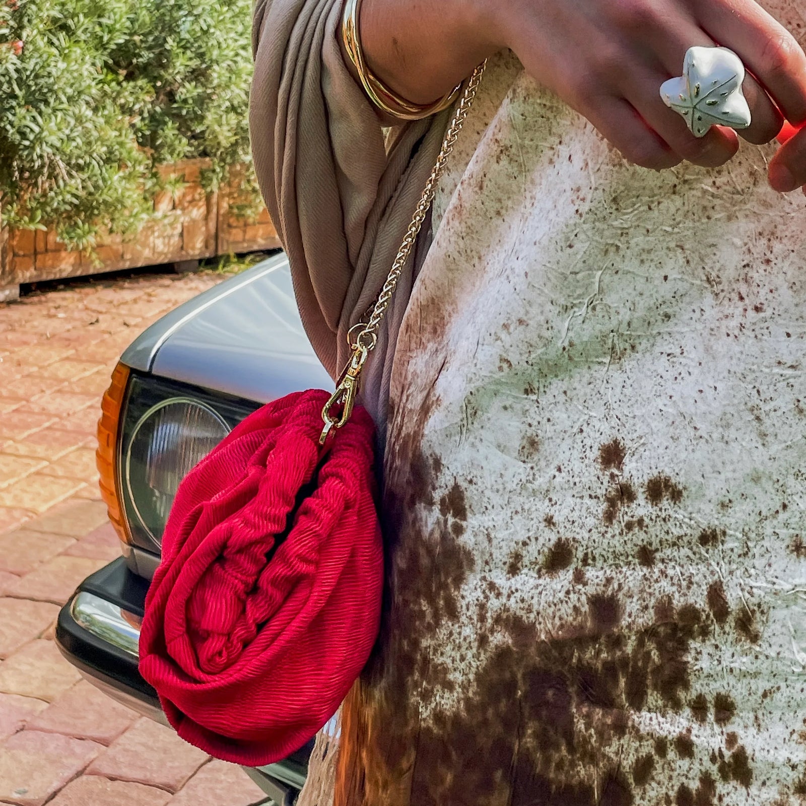 Red clutch bag with gold chain held by a hand against a car and stone wall background at a wedding.