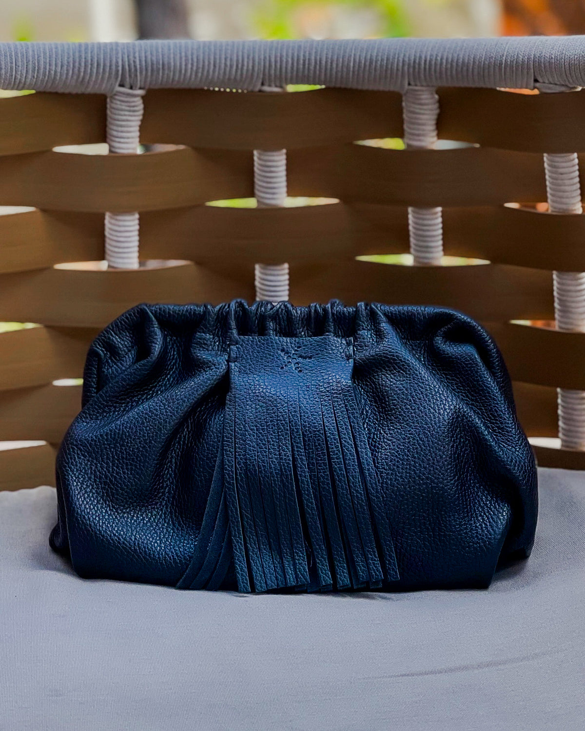 Black Natalia - Full Grain Leather Clutch Bag on a light gray surface with a wicker chair in the background