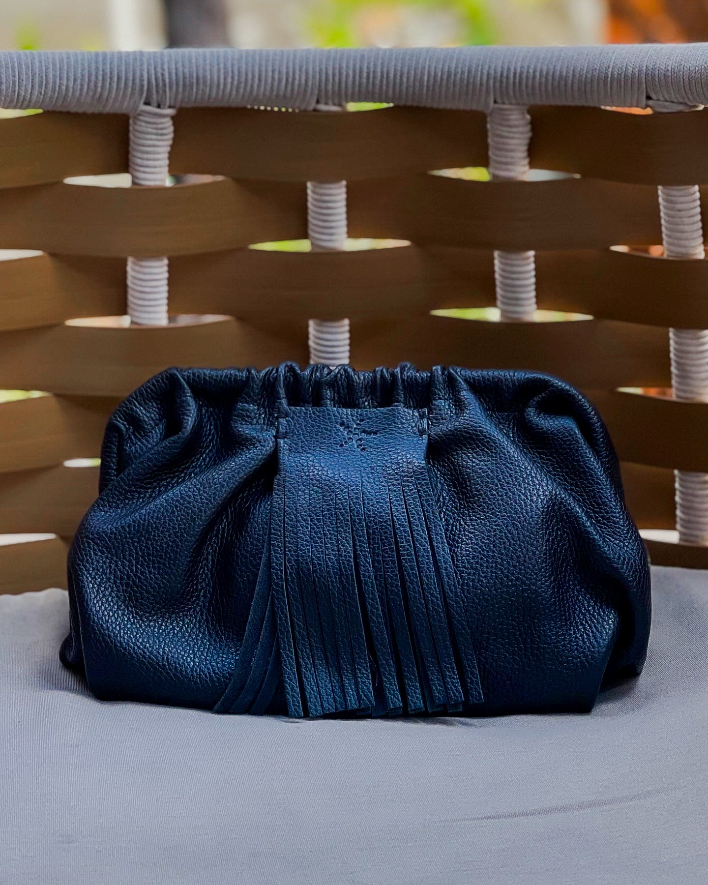 Black Natalia - Full Grain Leather Clutch Bag on a light gray surface with a wicker chair in the background