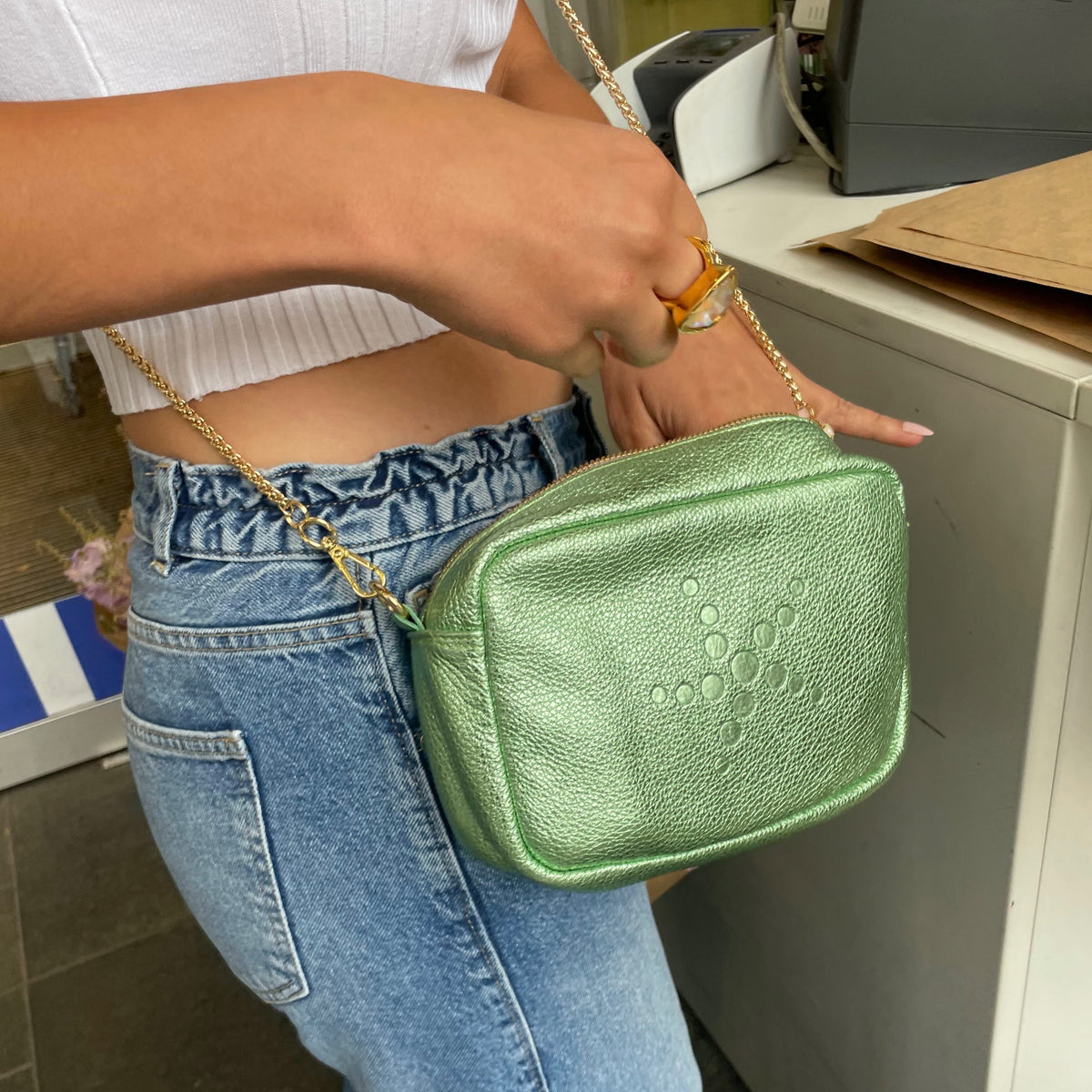 Person holding a Green Metallic Gabriela handbag with a chain strap in an indoor setting