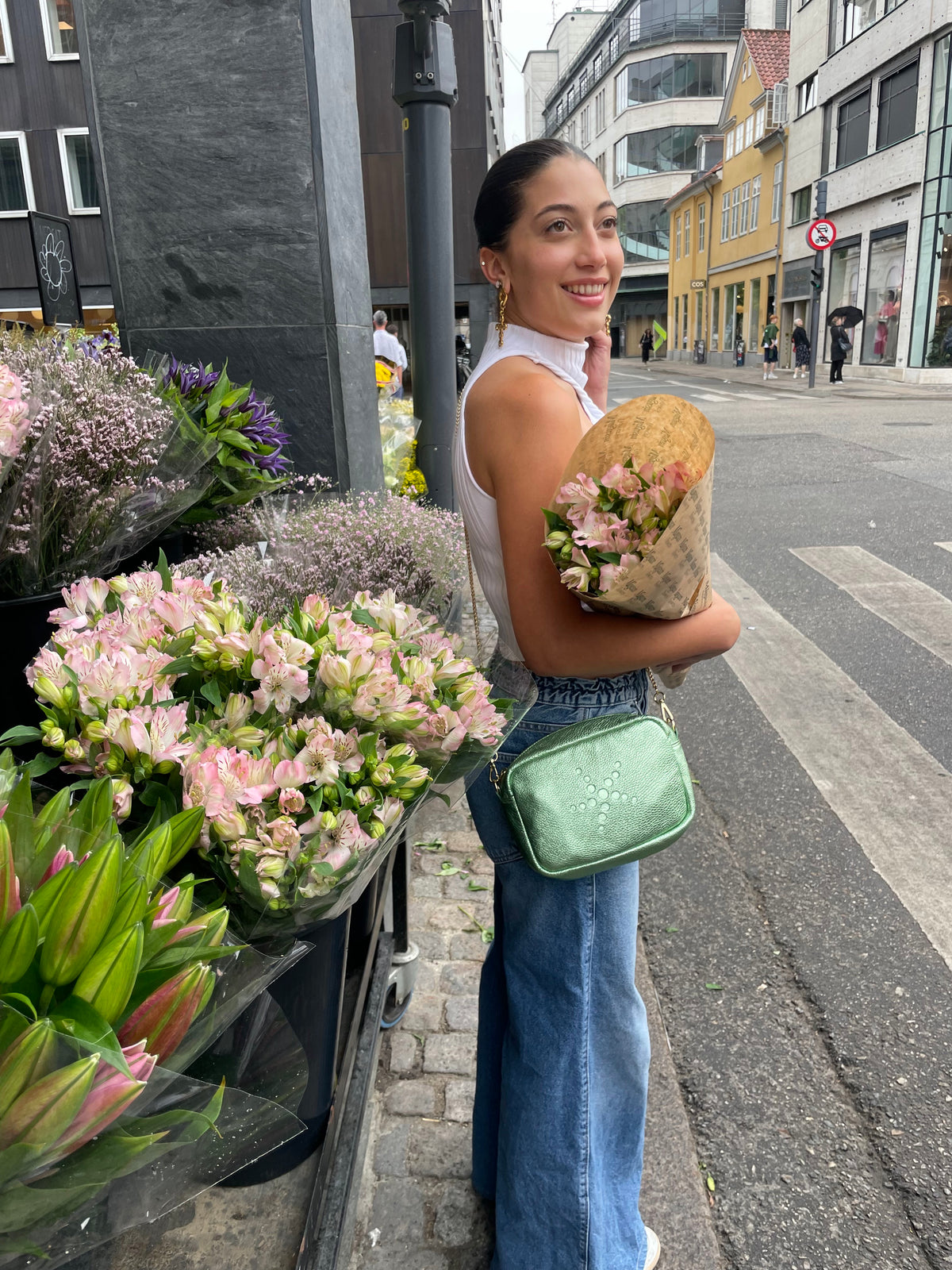 A woman holding a bouquet of flowers and wearing Green Metallic Gabriela on her shoulder walks down a street with flower stalls.