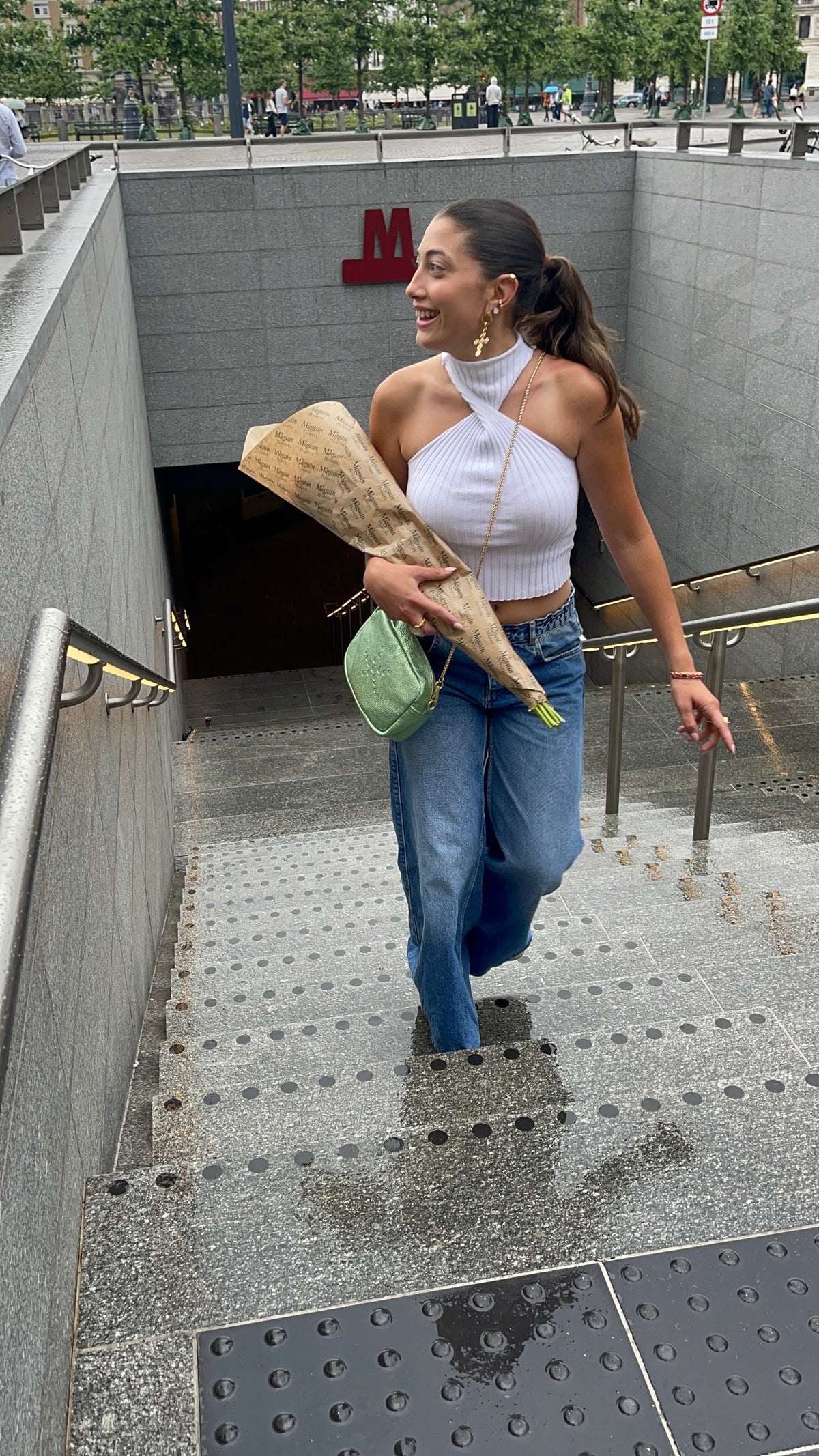 A woman walking out of a subway station holding flowers and a Green Metallic Gabriela bag.