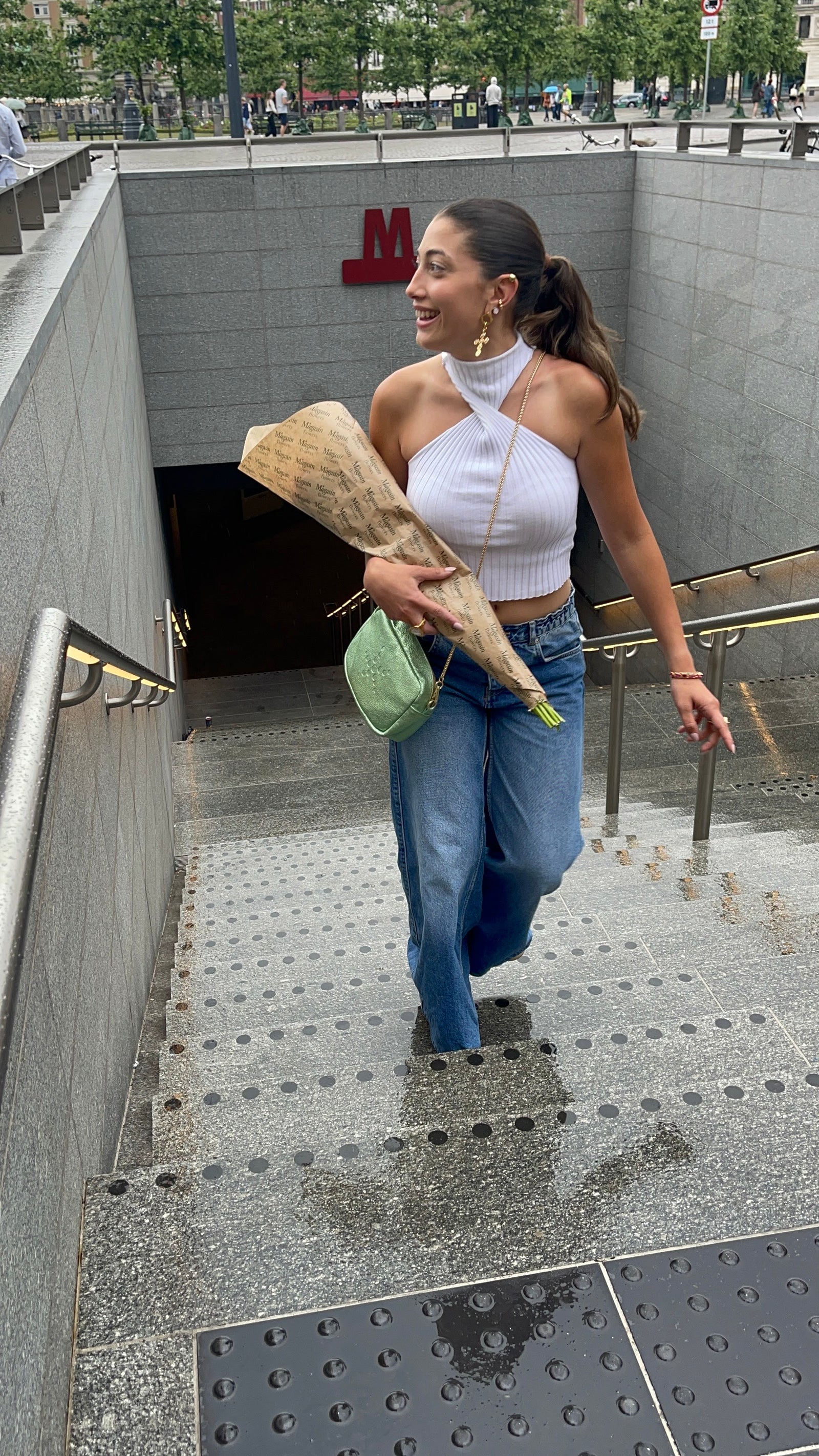 A woman walking out of a subway station holding flowers and a Green Metallic Gabriela bag.