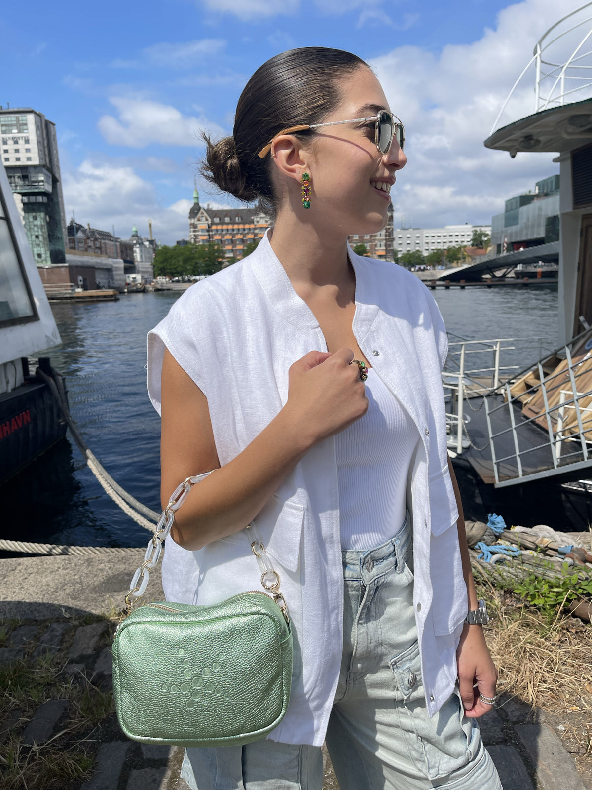 Close up Person standing by a waterfront with a Green Metallic Grabriela handbag on hand, boats, and buildings in the background