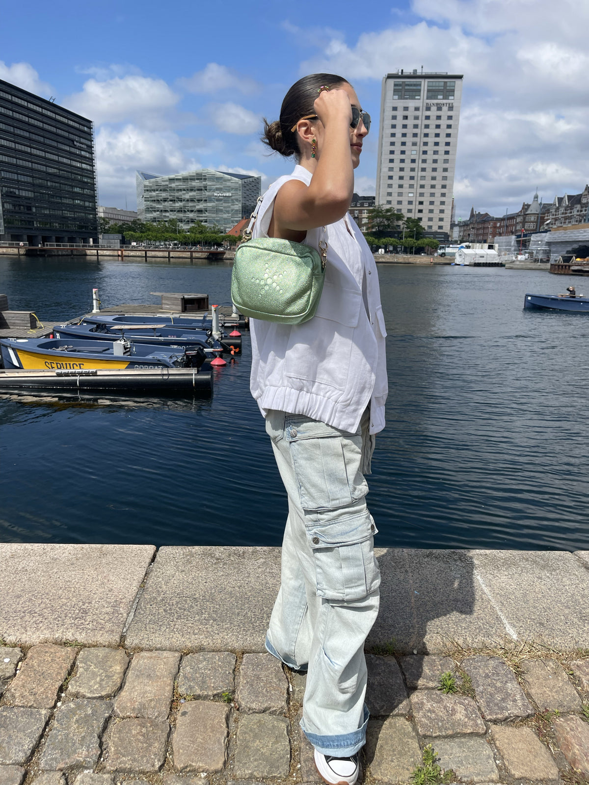 Person standing by a waterfront with a Green Metallic Grabriela handbag, boats, and buildings in the background
