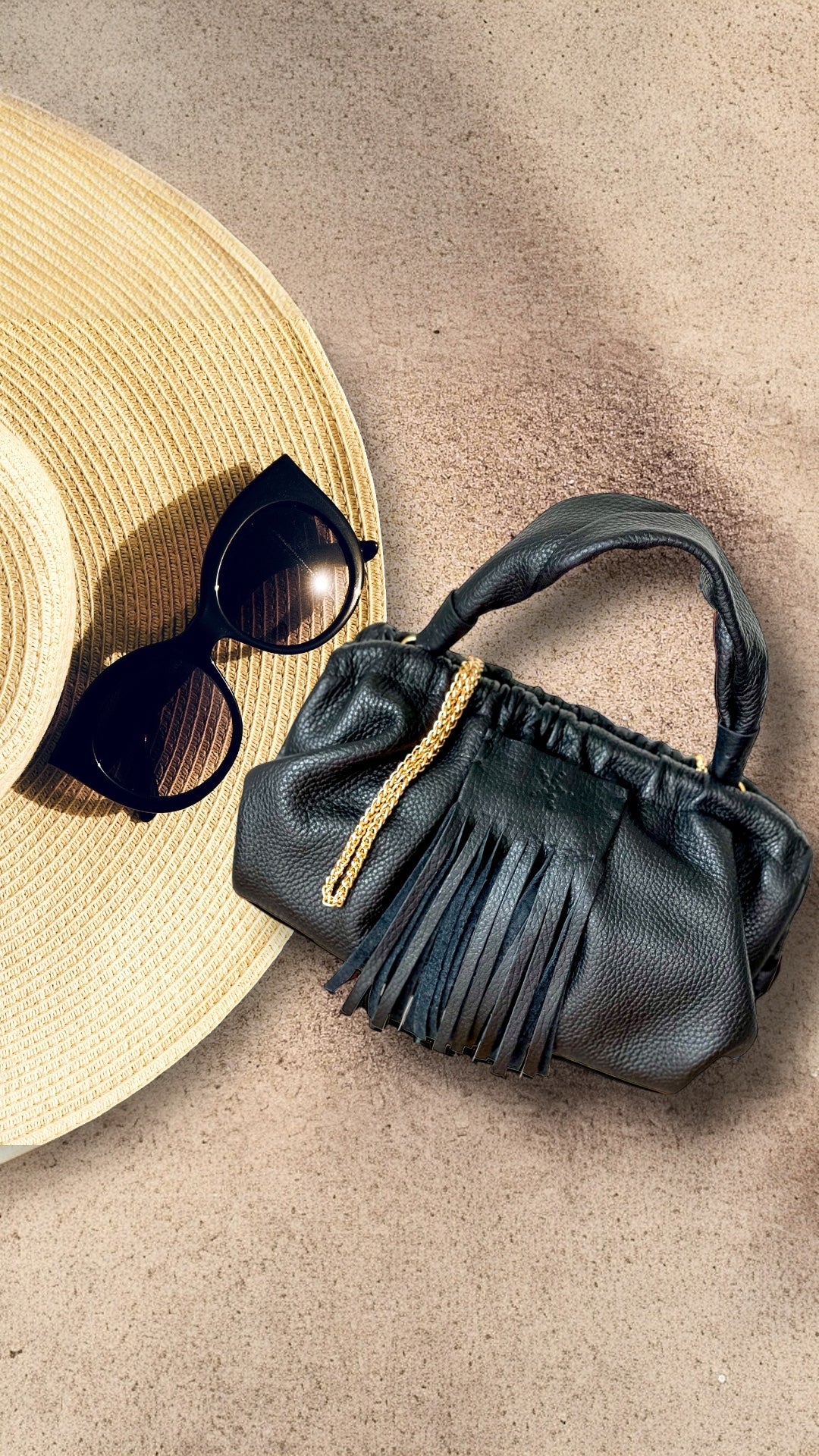 Black Natalia - Full Grain Leather Clutch  handbag with fringe details and sunglasses on a beige surface